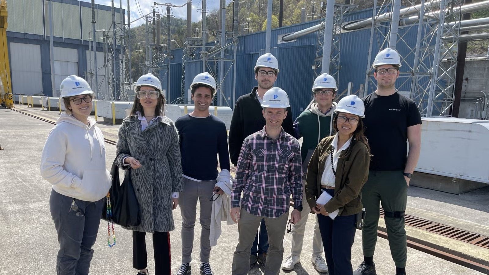 Students standing together outside plant facility, near fence, in casualwear and white hardhats.