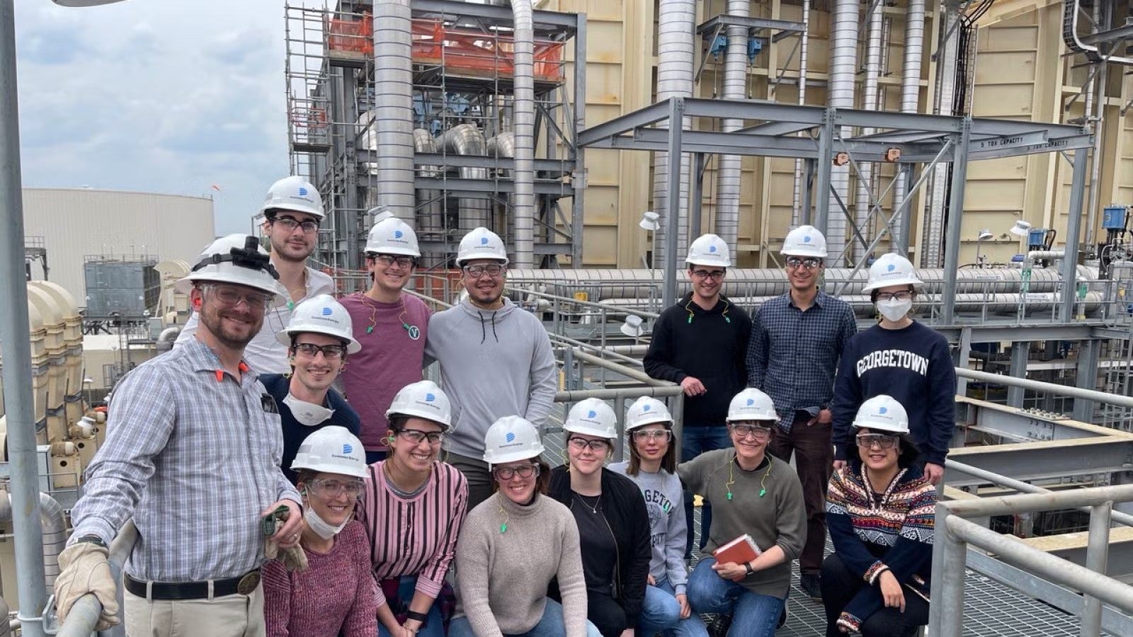 Group of students in casualwear and white hardhats visiting outside facility of power plant.
