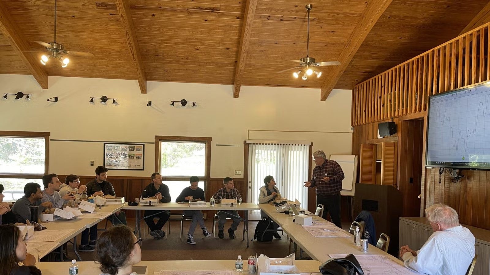 Students spread out, sitting at white 8-foot tables connected in a large rectangle while someone speaks at front of room.