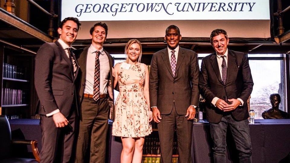 Ignac poses with four other people on a stage with the words "Georgetown University" displayed on a screen
