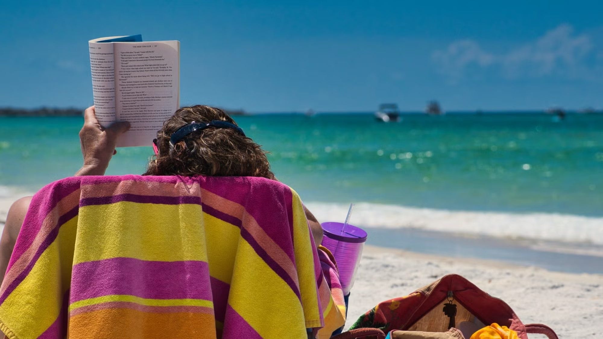 Person sitting in chair with pink, yellow, and orange-striped towel hanging over the back, reading, with the ocean in the background.
