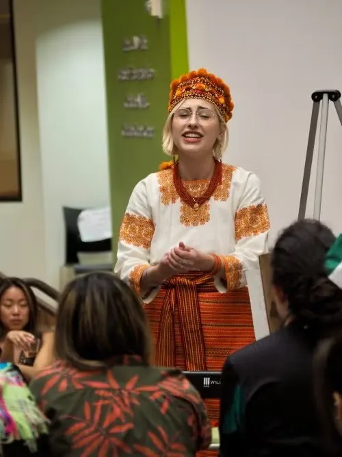 Sophia Farion dressed in orange and white traditional Ukrainian attire while standing in front of classroom, with students listening to her presentation.