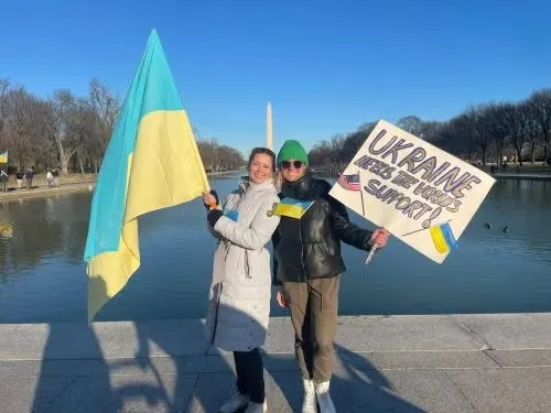 Marta holding Ukraine flag on National Mall, in front of Washington Monument, with student holding handwritten sign for Ukrainian support.