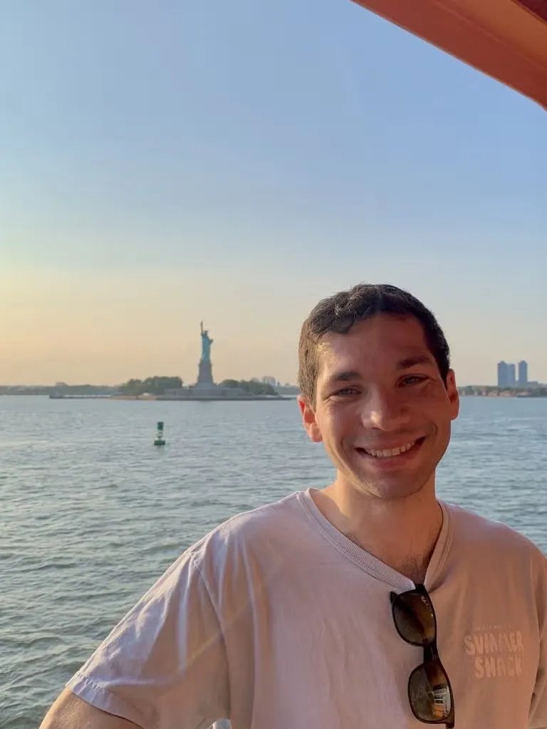 Caleb Diamond standing with Statue of Liberty in background, wearing a light gray t-shirt.