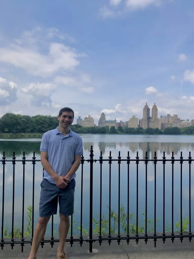 Caleb Diamond standing in front of lake with New York City in the background, wearing a light blue polo shirt and navy blue chino shorts.