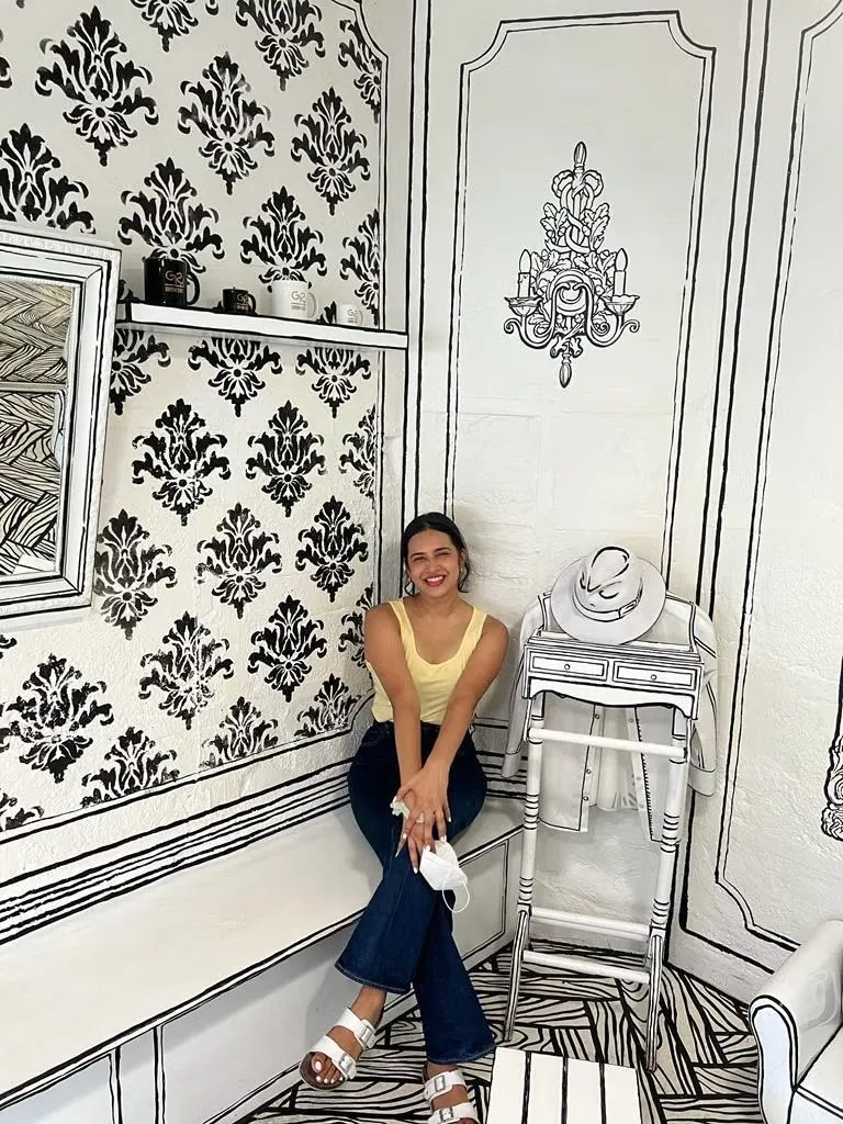 Anaga Krishna wearing yellow tank top and dark denim jeans with white sandals sitting on bench in white room with black pattern.