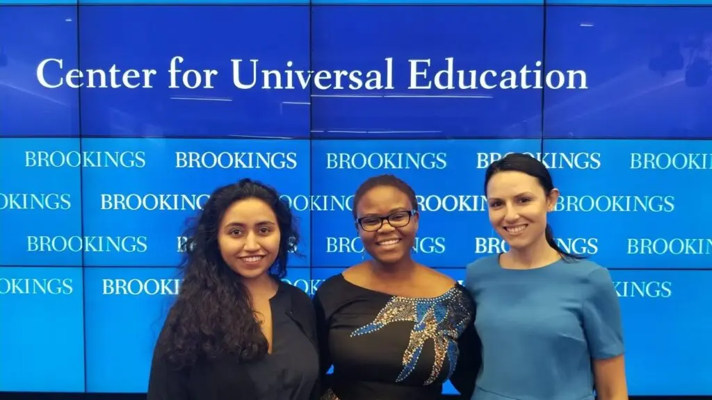 Three young women standing in front of blue and white Center for Universal Education sign.
