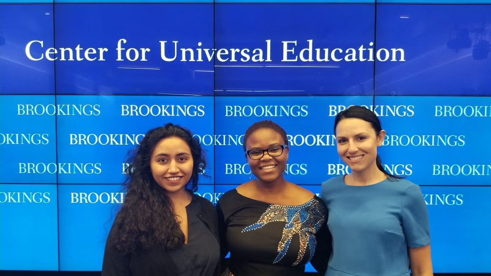 Three young women standing in front of blue and white Center for Universal Education sign.