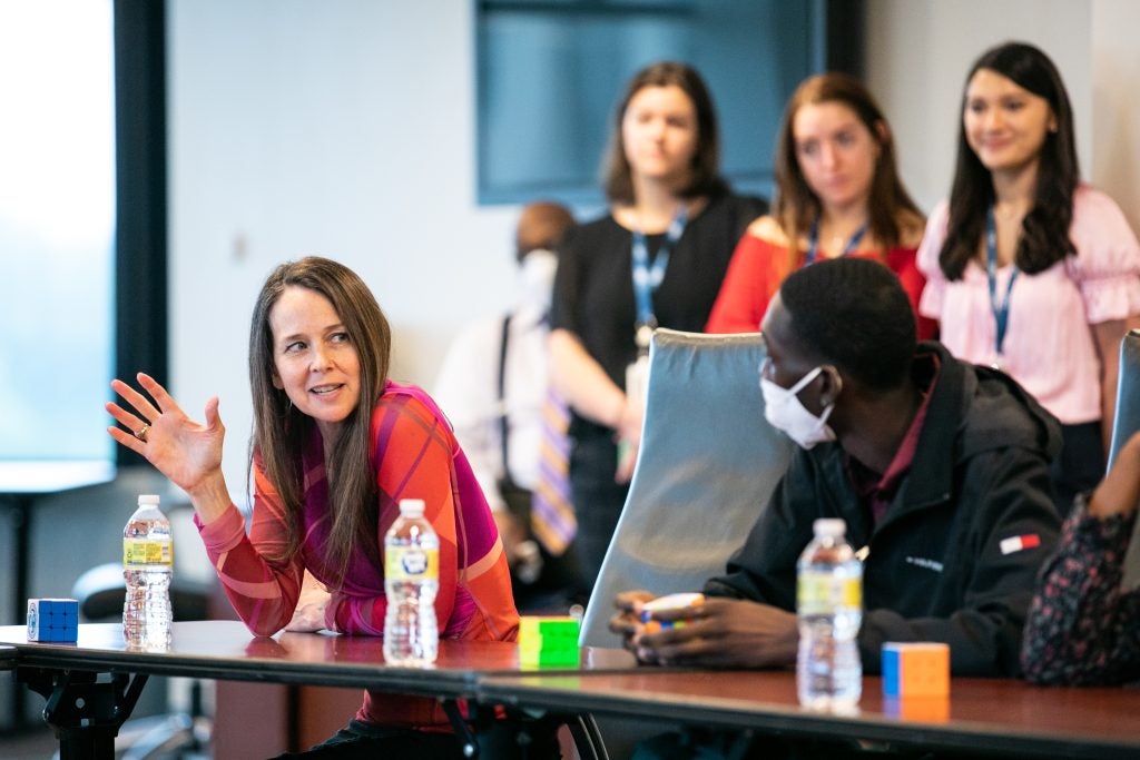 Jen Easterly gestures with right hand while speaking, seated at a table. Three staff members stand to right behind her.