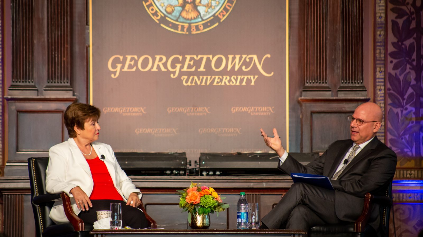 Dean Hellman gestures with his hands as he asks Georgieva a question. The two are seated side-by-side while on-stage in Gaston Hall.