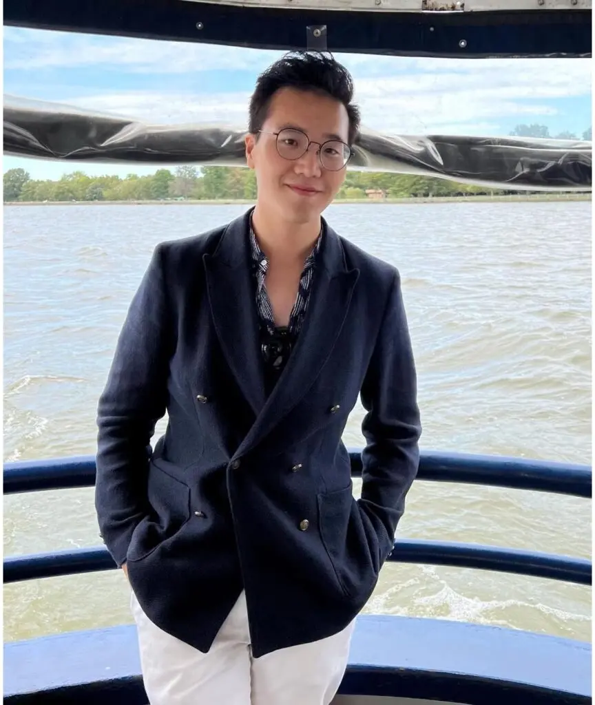 Alex Chou Headshot, wearing black-rimmed glasses, a navy blue blazer, navy shirt, and white pants.