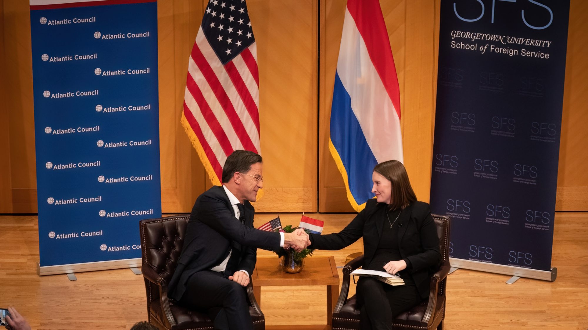 Mark Rutte and Amy Mackinnon shake hands while on-stage, with the American and Dutch flags in the background.