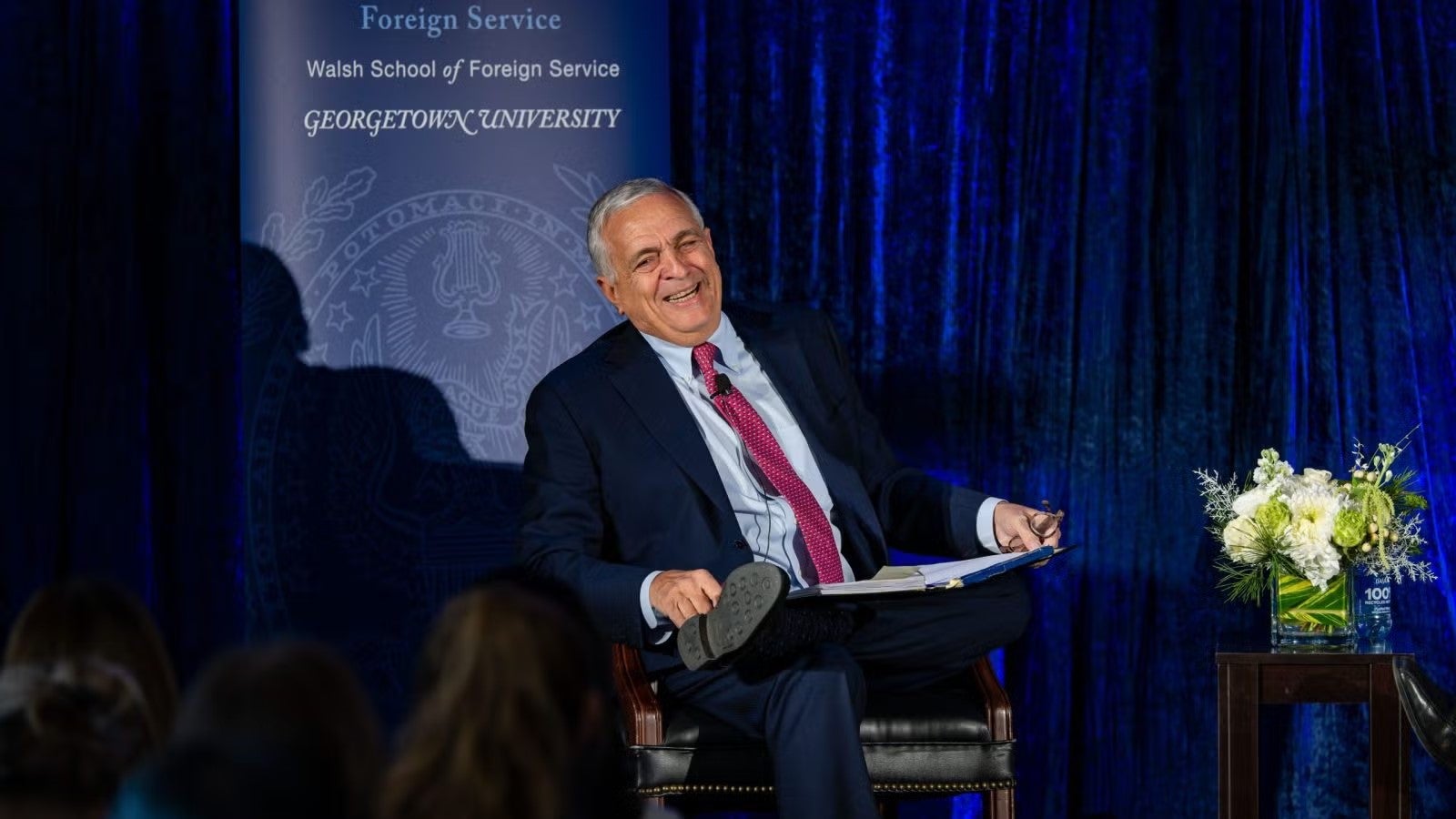 George Tenet sitting on stage at SFS 100 year gala wearing navy blue suit, blue shirt, and red tie.