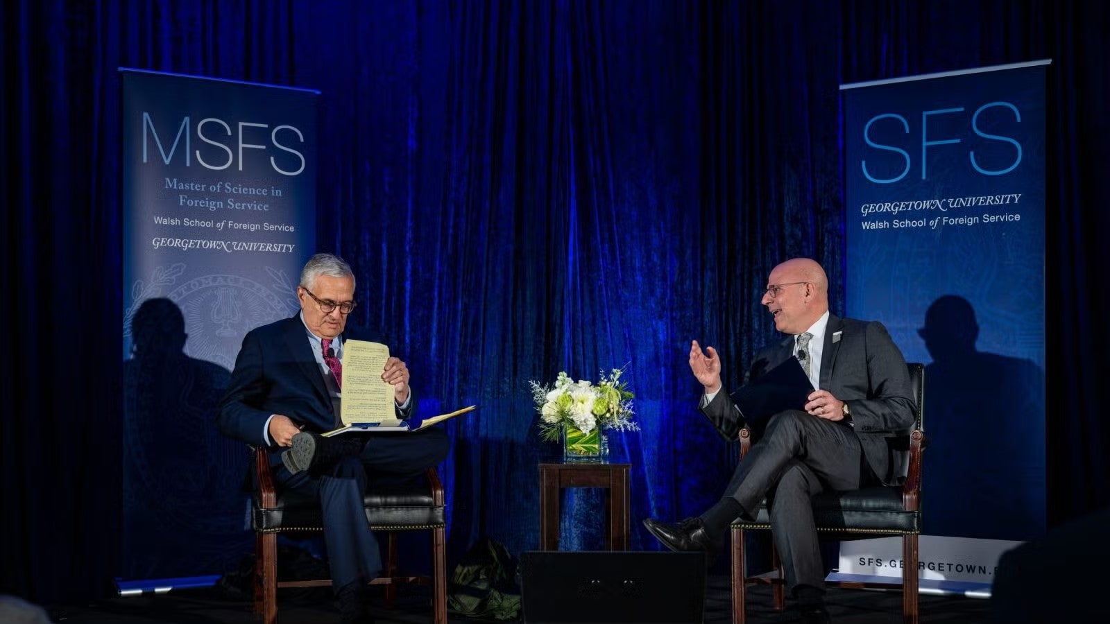 George Tenet and Joel Hellman sitting on stage at 100 year gala, talking with each other.