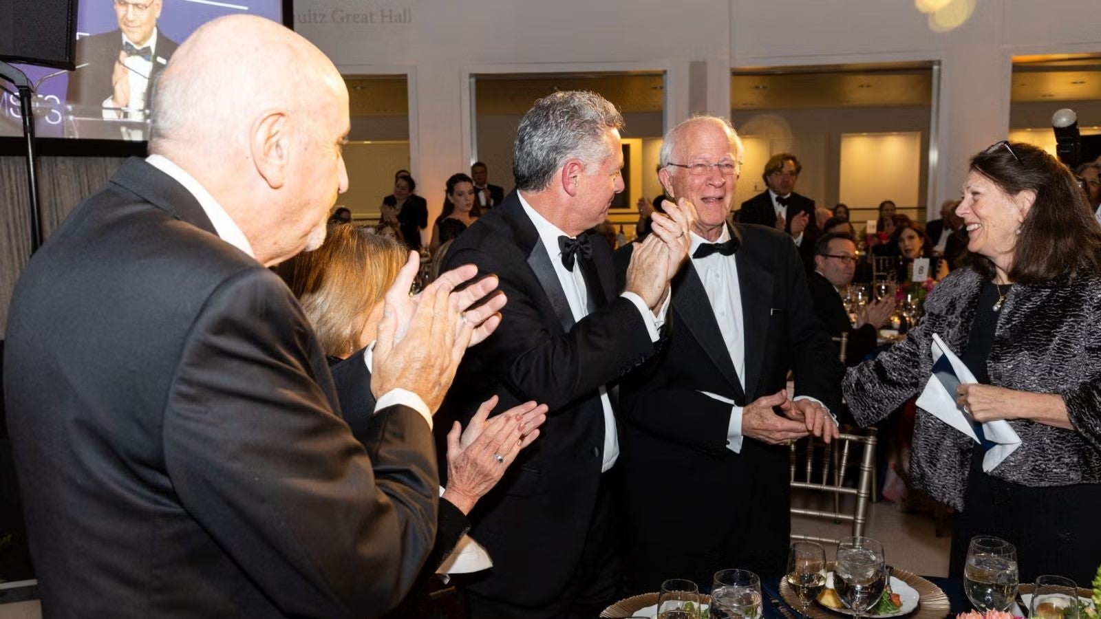 Attendees in dark formalwear clapping at gala.