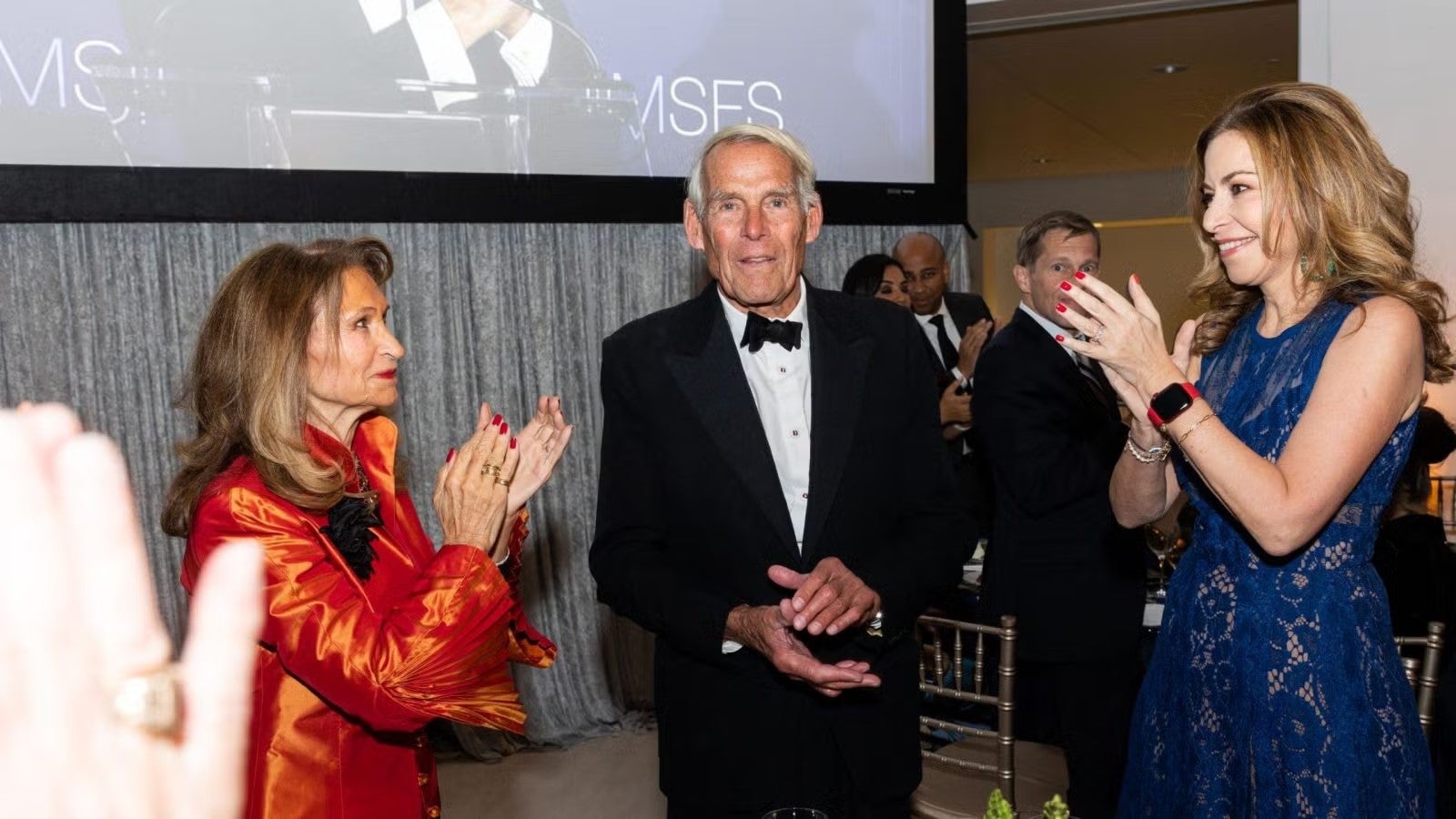 Two women and a man, in formalwear, clapping  at gala.