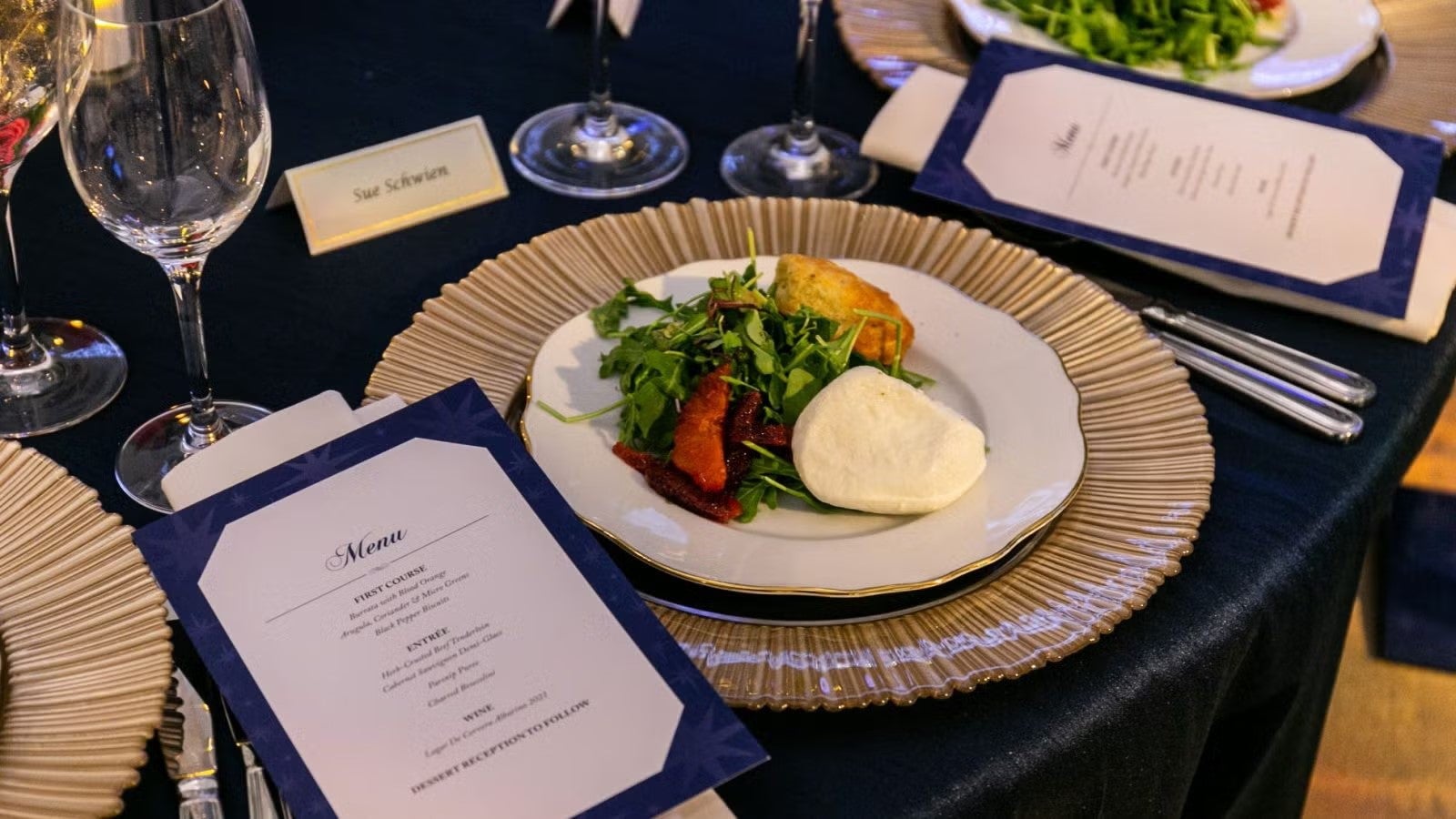 Dinner plate at gala, placed on table next to dinner menu, featuring mozzarella, spring greens, blood oranges, and a dinner roll.