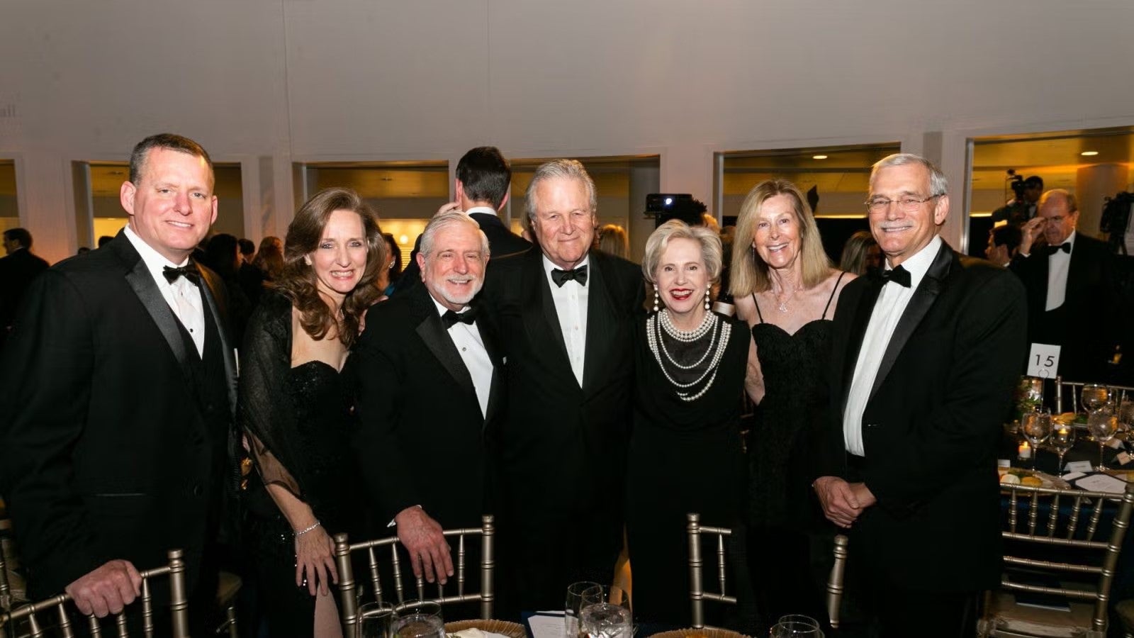 Six men and women dressed in formalwear standing around a formal, round dinner table at gala.