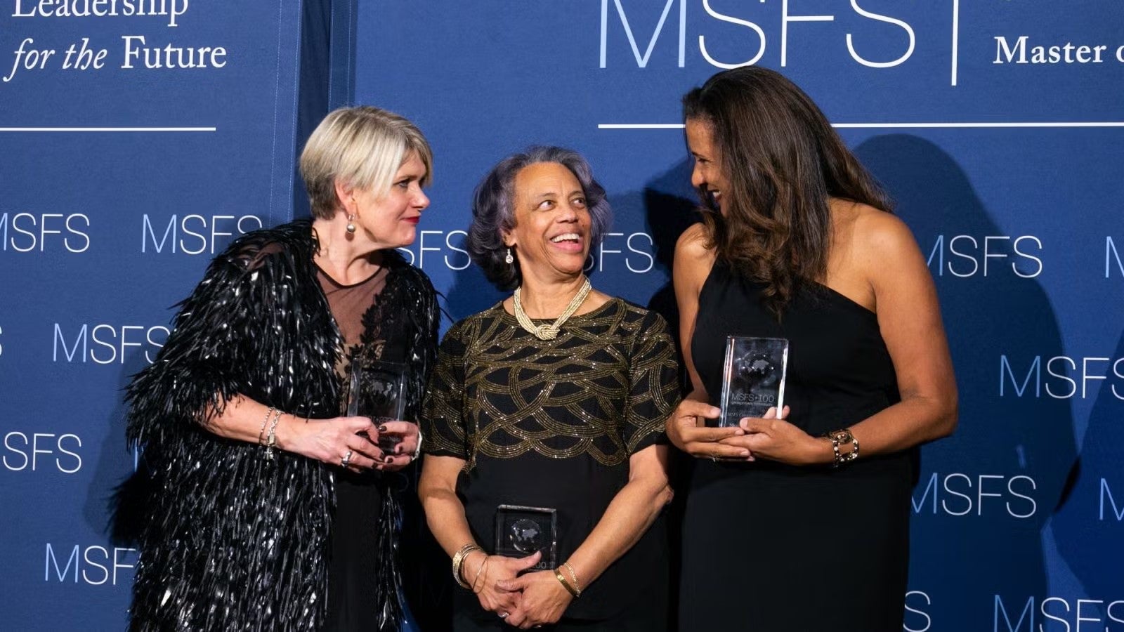 Three women in dark formalwear standing together with awards at gala.