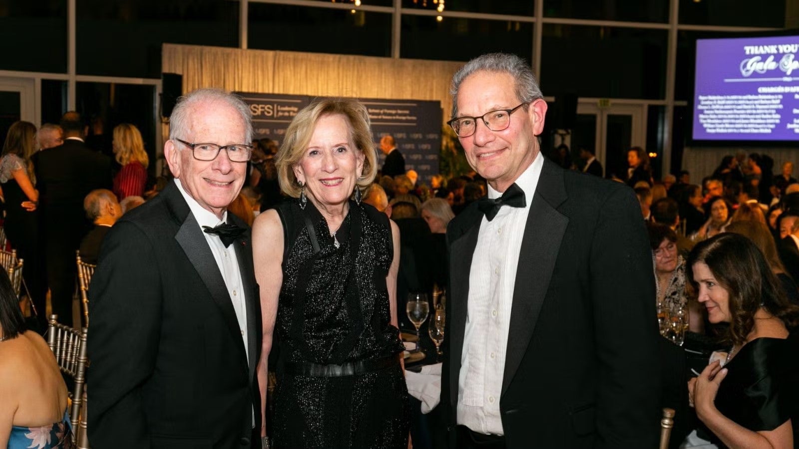 Two older men and one woman standing together in formalwear, posing for photo at gala.