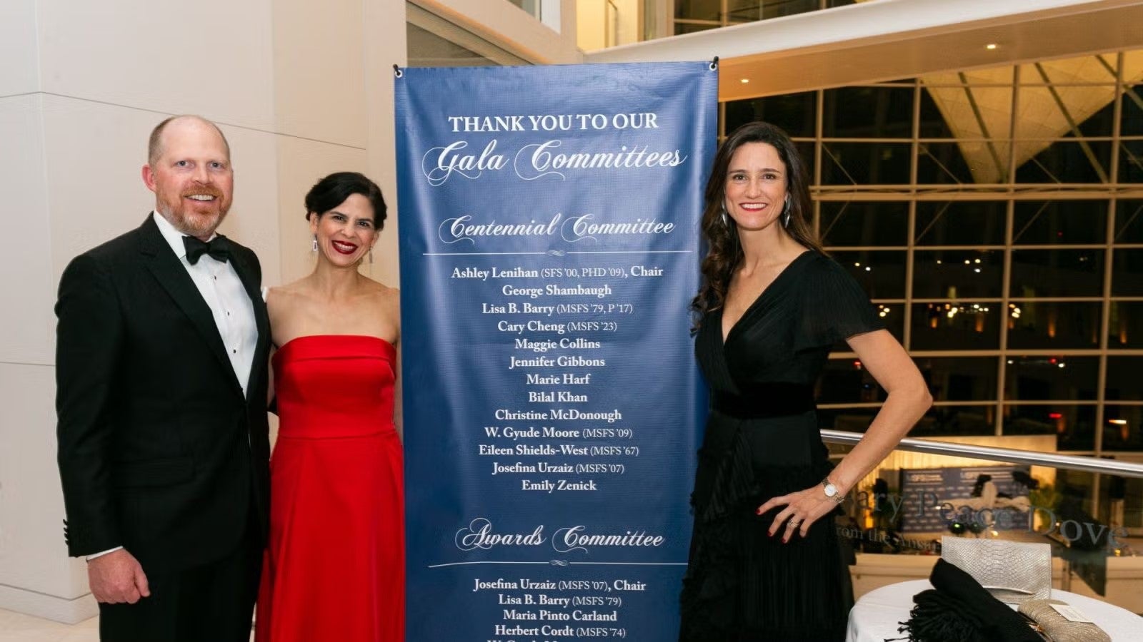 Two women and a man from the gala committee, in formalwear, standing next to gala banner.