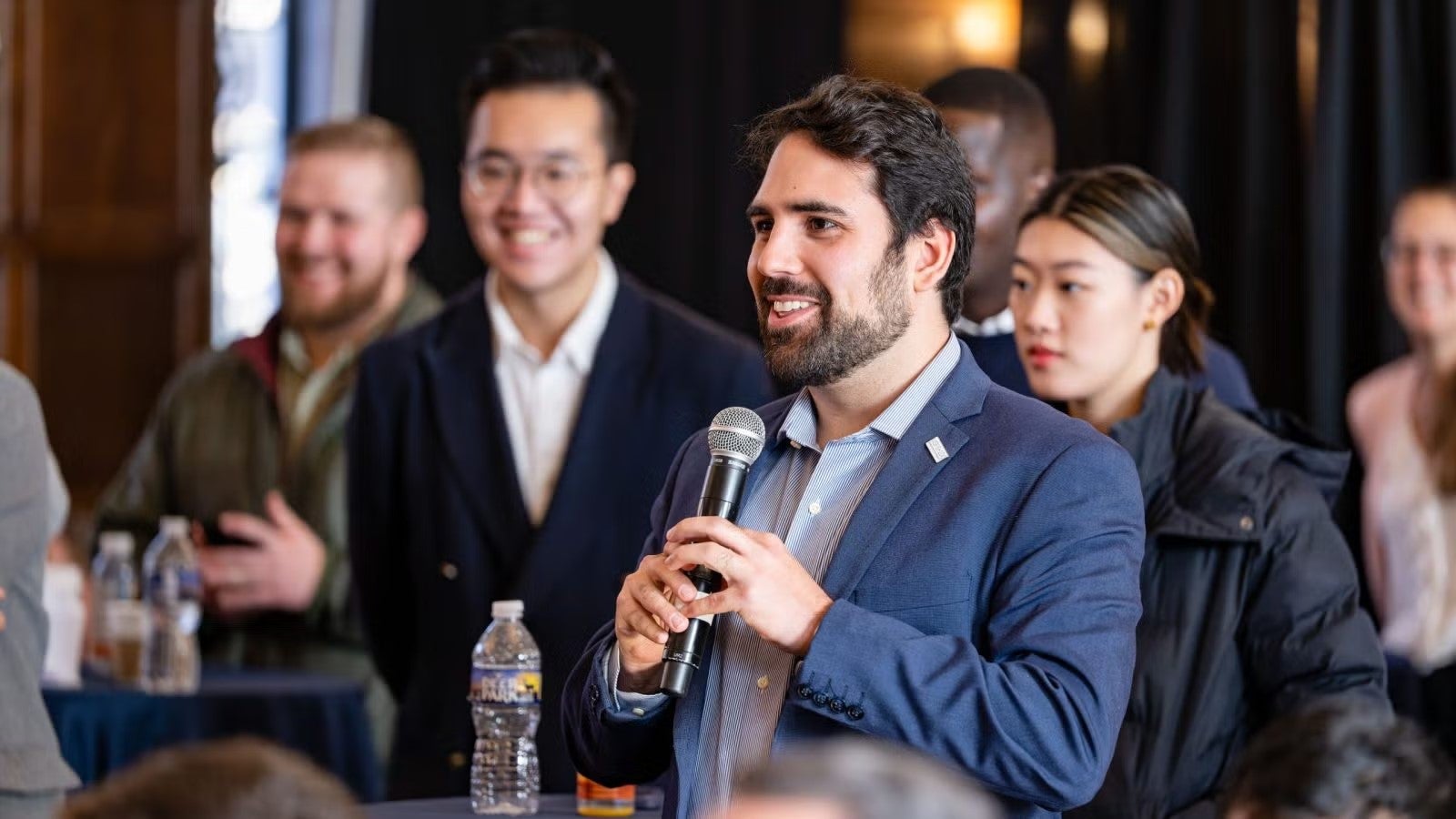Young man with dark hair and dark beard, wearing navy blue blazer and blue shirt stands in middle of room, holding microphone.