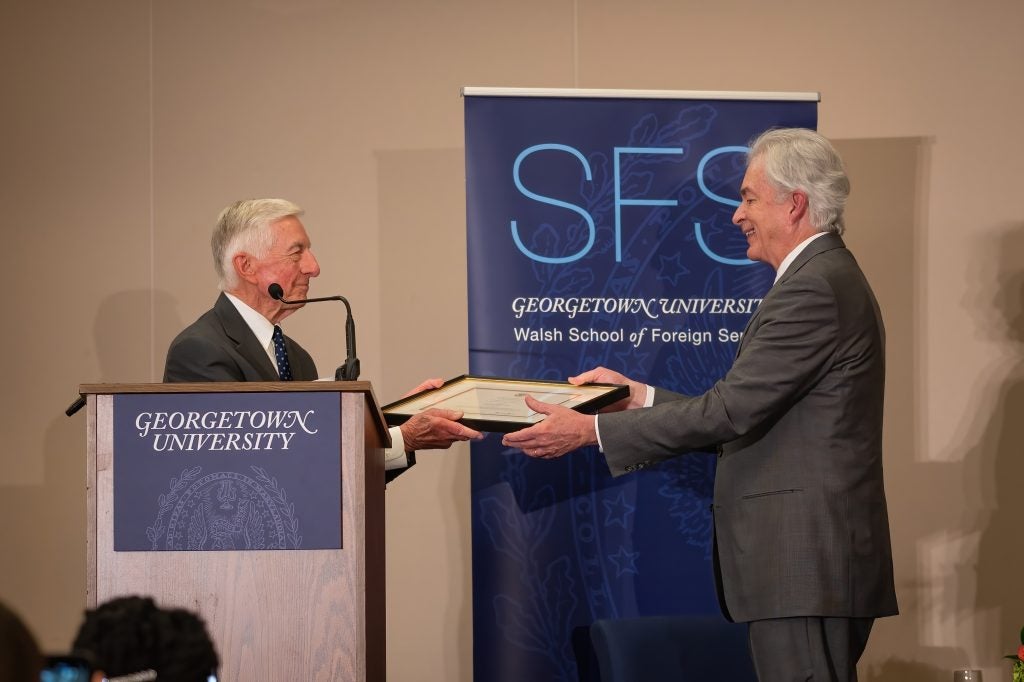 Hogan presents Burns with the Trainor Award. The two men stand, smiling, side-by-side on stage with both of their hands on the plaque.