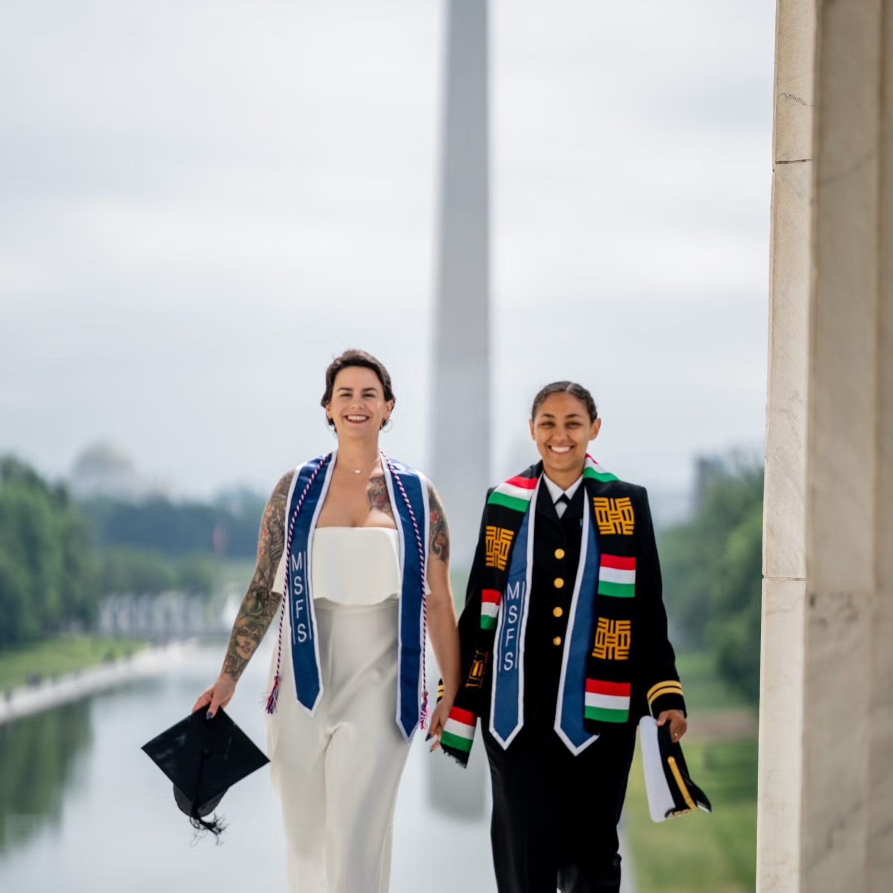 Jasmin Alsaied photographed on National Mall, in front of Washington Monument, wearing formal military dress, commencement stole, and holding military cap, standing, holding hand of another SFS student, a young female with short, dark hair, wearing white jumpsuit, commencement stole, and holding graduation cap.