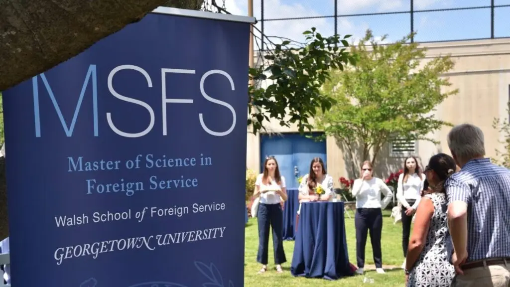 A blue banner for MSFS at the Georgetown Observatory welcoming SFS alumni  while a few young women stand nearby looking on towards a woman speaking at the event.