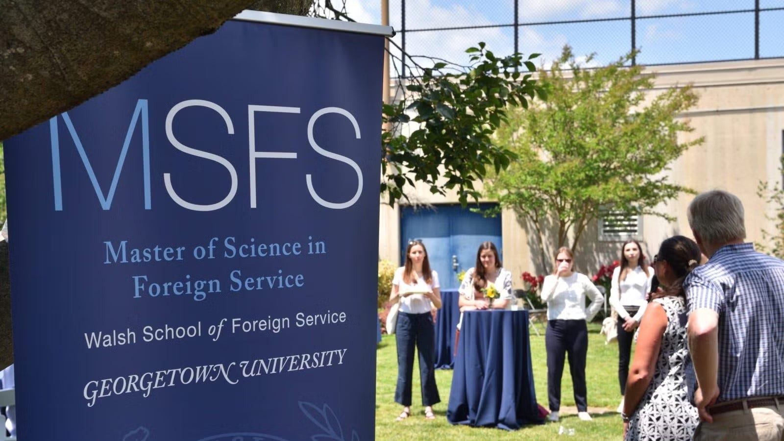 A blue banner for MSFS at the Georgetown Observatory welcoming SFS alumni while a few young women stand nearby looking on towards a woman speaking at the event.