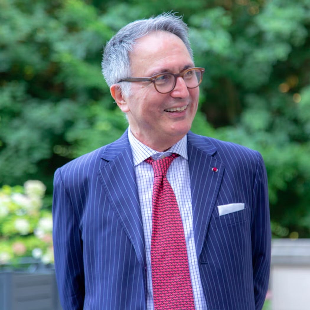 Raoul Delcorde Headshot in Navy blazer, light blue shirt, and red tie.