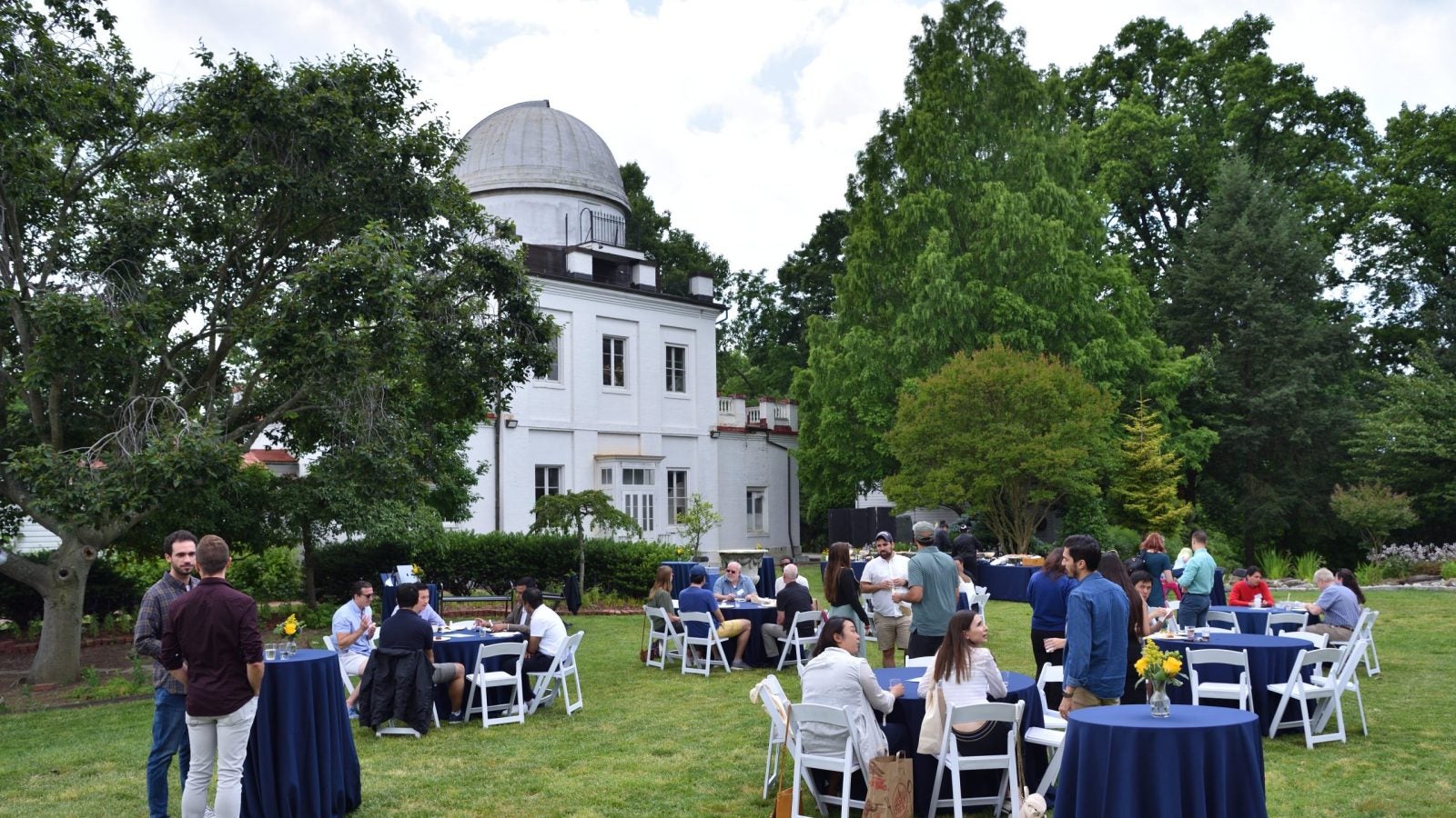 Three tables with people seated at them outside near a larch white brick building.