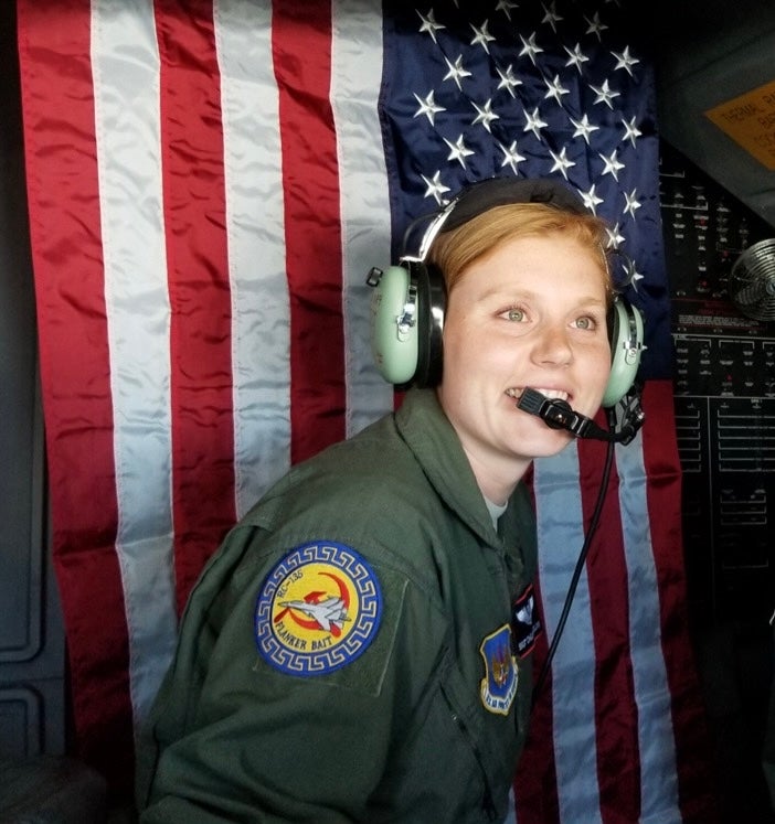 Person in a flight suit with a headset, smiling, in front of an American flag inside an aircraft.