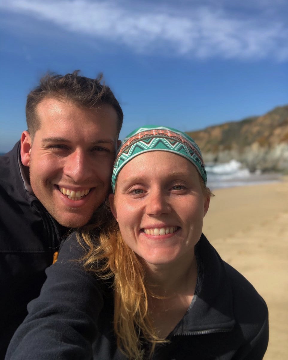 Two smiling individuals taking a selfie on a sunny beach with cliffs in the background.
