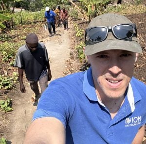 A selfie of Evan Anthony wearing a blue IOM shirt standing in front of storm damage in Burundi.