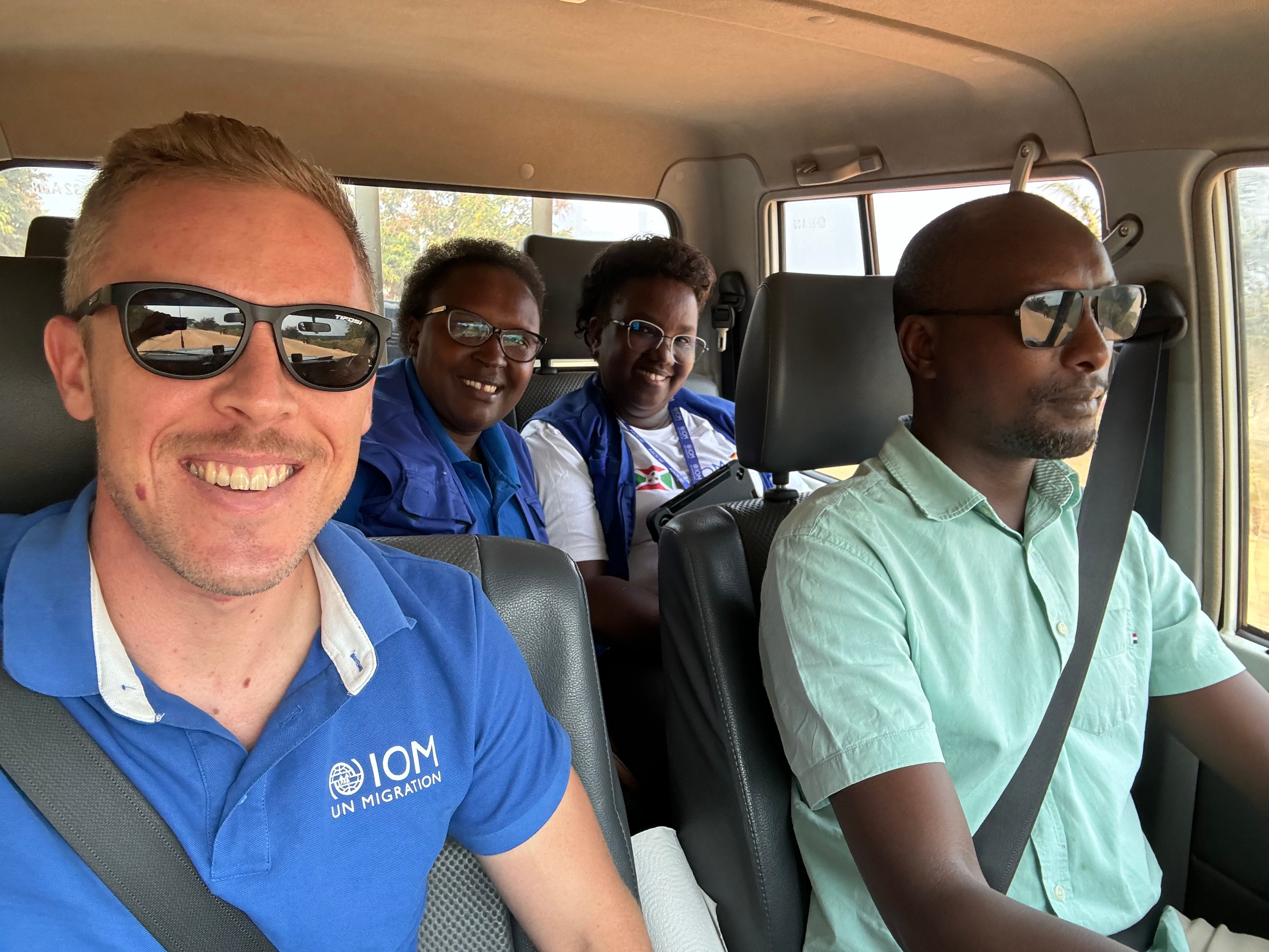 Four individuals in a vehicle, smiling; one is wearing a blue shirt with the IOM (International Organization for Migration) logo.