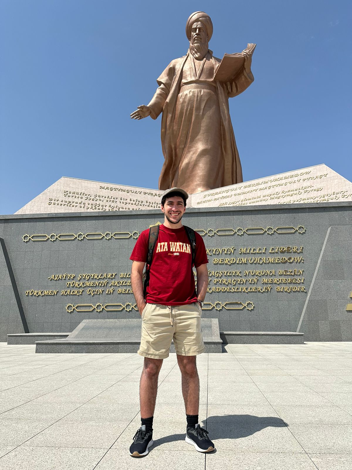 Person wearing red t-shirt and khaki shorts smiling in front of the bronze statue of Magtymguly Pyragy.