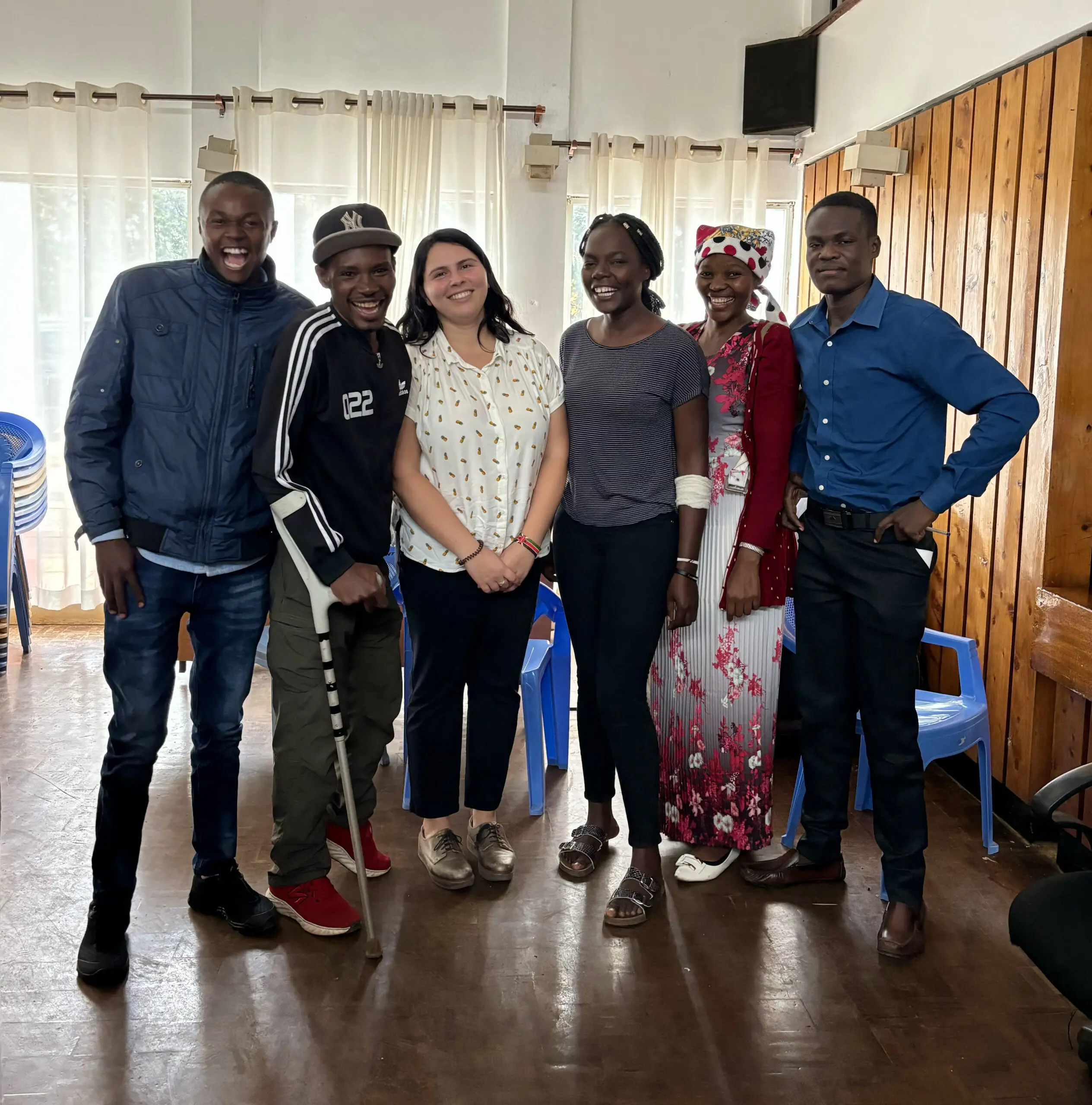 Six individuals posing with smiles in a room with wooden walls and a window. They are dressed in a mix of casual and traditional attire.
