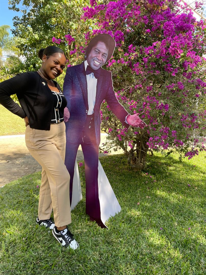 A person smiling and standing next to a life-sized cardboard cutout of James Brown in a garden with a blooming pink bougainvillaea bush in the background.