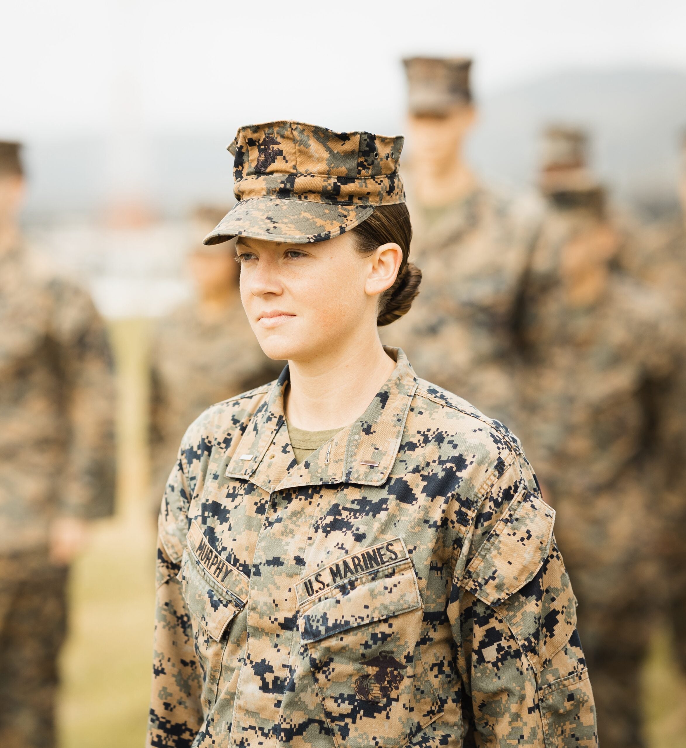 A U.S. Marine in camouflage uniform standing solemnly against a backdrop of fellow Marines slightly out of focus.