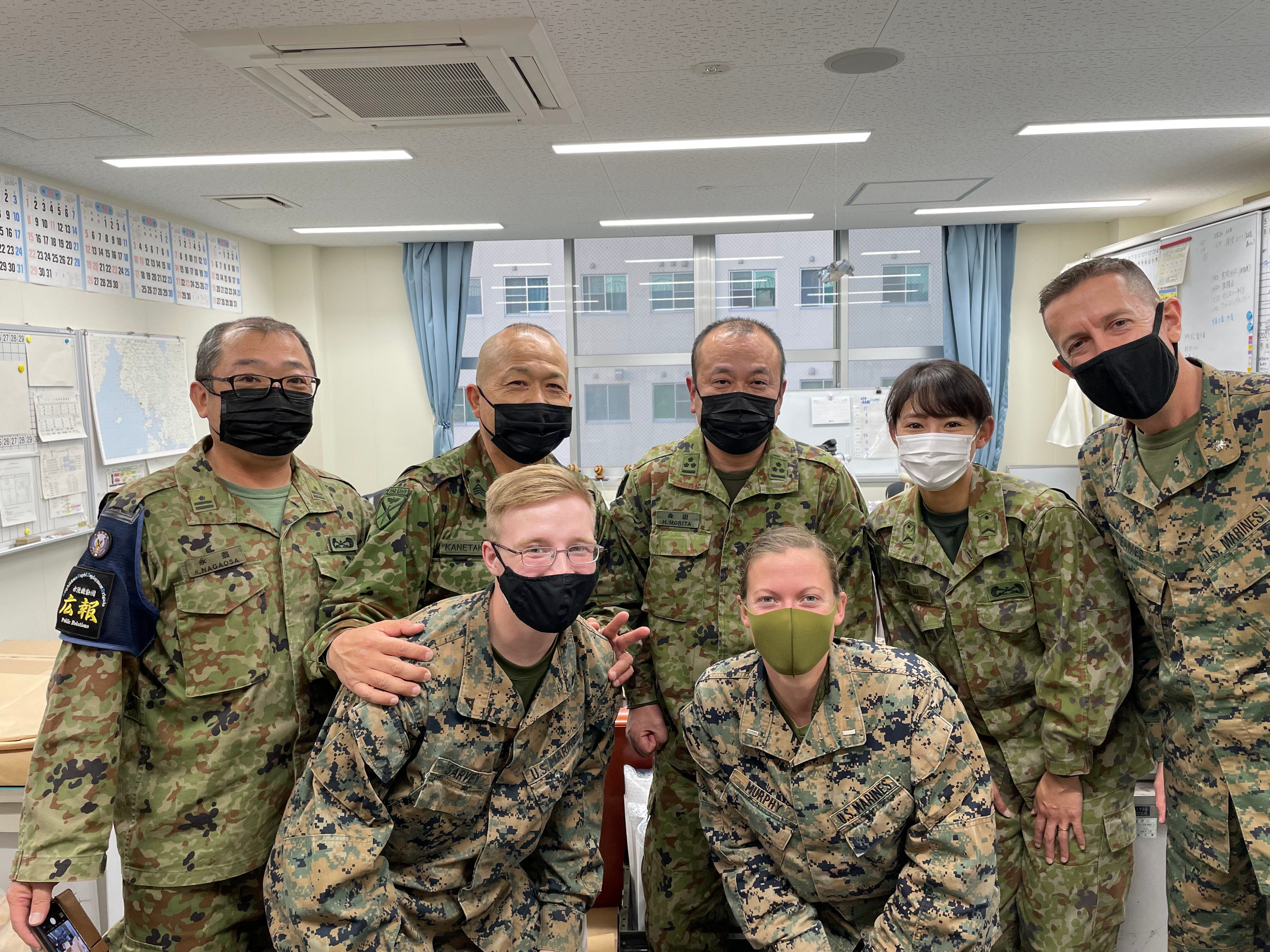 Group of eight military personnel in camouflage uniforms, wearing masks, posing jovially in an office setting with maps on the wall behind them.