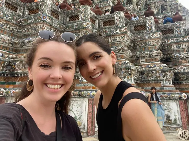 Two smiling women wearing black tops taking a selfie with the ornate Wat Arun temple in the background.