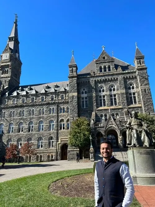 A person smiling in front of the historic Healy Hall at Georgetown University on a sunny day.