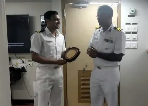 Two naval officers in white uniforms smiling and exchanging a plaque inside a ship cabin.