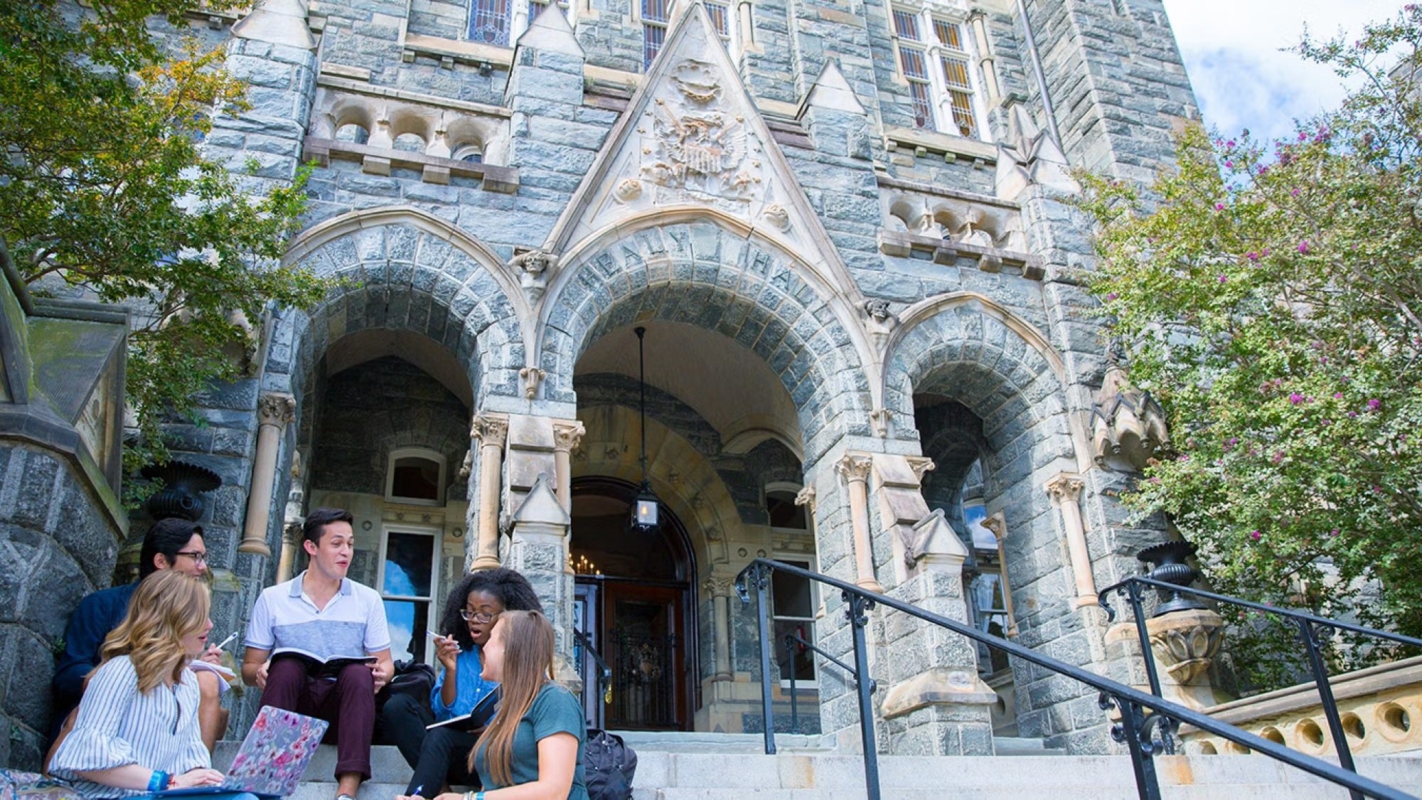 Five students sitting and discussing with notebooks in front of the ornate entrance of Georgetown University's Healy Hall.