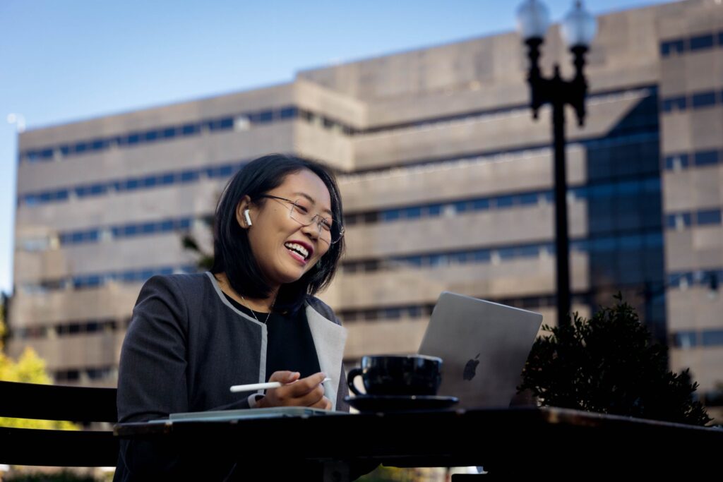 Student in a navy blazer smiling and typing on a laptop outside.