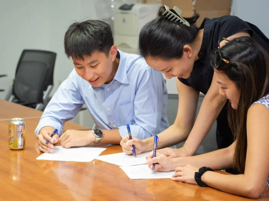 Three students at a table, looking at a paper together and smiling.