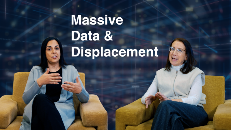 Two people seated in armchairs are engaged in a discussion in front of a digital background that reads &quot;Massive Data &amp; Displacement.&quot;