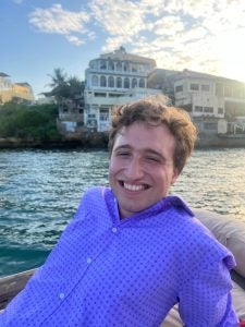 Person smiling on a boat with a scenic view of historic coastal buildings in the background under a clear sky.