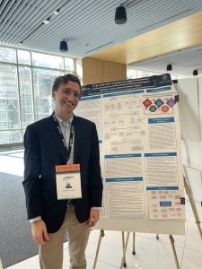 A person presenting a poster at a conference, smiling and wearing a conference badge. The poster contains various diagrams and text sections.